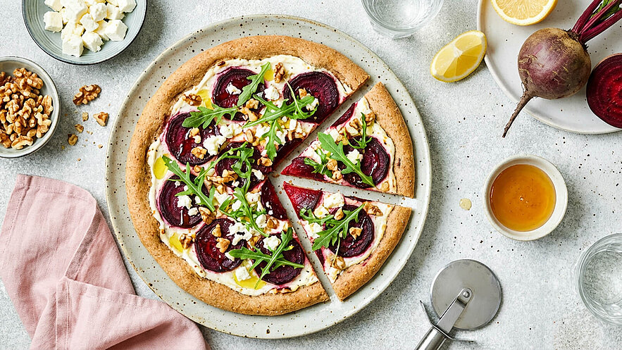 Eine runde Pizza mit einem Boden aus Vollkorn, belegt mit Rote Bete, Feta, Walnüssen und Rucola, die in acht Stücke geschnitten ist. Neben der Pizza liegen eine rosa Serviette, ein Gläser, Zitronenscheiben und eine Rote Beete.