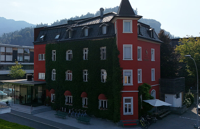 Ein rotes Gebäude mit mit Grün bewachsenen Wänden steht im Vordergrund. Im Hintergrund sind sanfte Hügel zu sehen. Vor dem Gebäude gibt es eine Treppe, Fahrradständer und einen Bereich mit kleinen Bäumen. Die Szene vermittelt eine ruhige, einladende Atmosphäre.