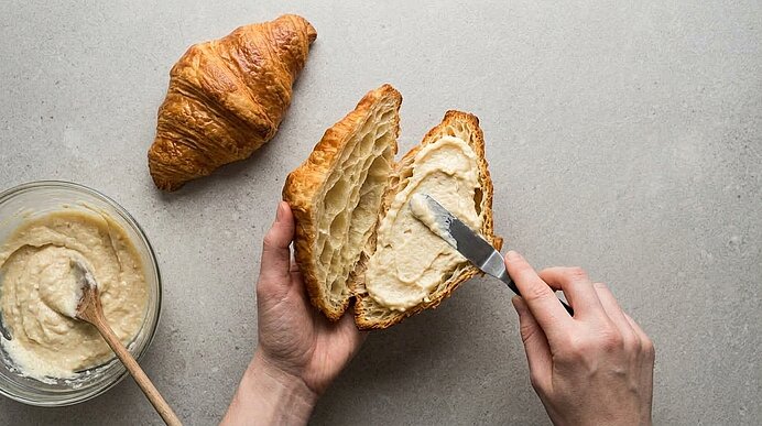 Zwei Hände streichen mit einem Messer Mandelcreme in ein aufgeschnittenes Croissant auf grauer Arbeitsplatte.