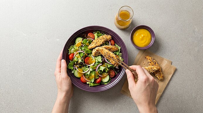 Hände halten eine Schüssel mit gemischtem Salat (Rucola, Gurke, Kirschtomaten, roten Zwiebeln) und knusprigen panierten Hähnchenstreifen; eine Gabel hebt einen Streifen. Daneben auf Papier weitere Streifen und ein Schälchen Senfdressing.