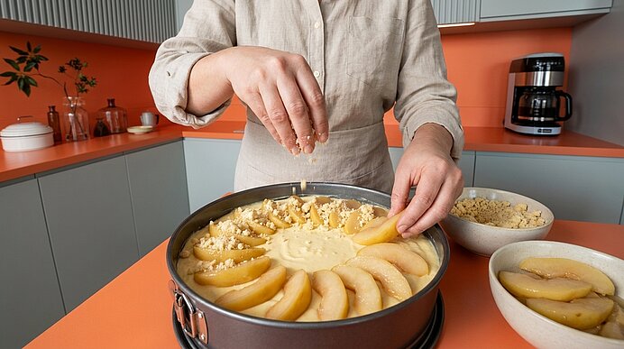 Hände einer Person legen Birnenscheiben in einen mit Teig gefüllten Springform-Kuchen und streuen Streusel darüber.