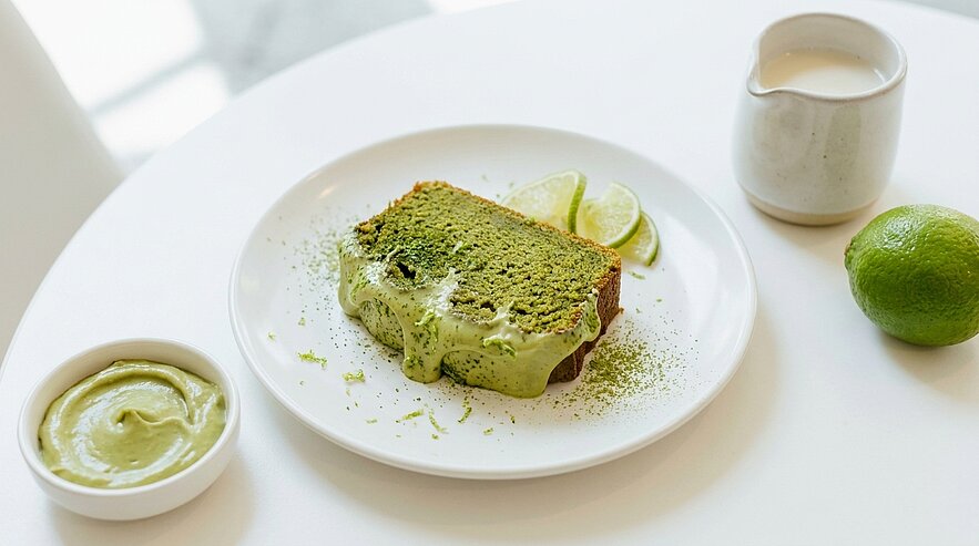 Scheibe grüner Kuchen mit verlaufender, hellgrüner Creme auf weißem Teller, dazu Limettenspalten und kleines Schälchen mit grüner Creme.