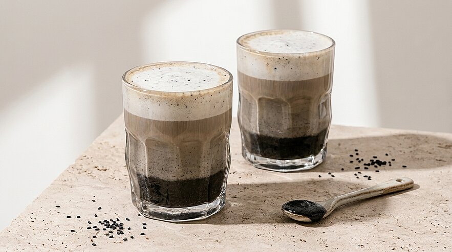 Zwei klare Gläser mit geschichtetem Kaffeegetränk — dunkle Basis, hellbraune Mittelschicht und cremiger Schaum oben; ein Löffel liegt daneben.