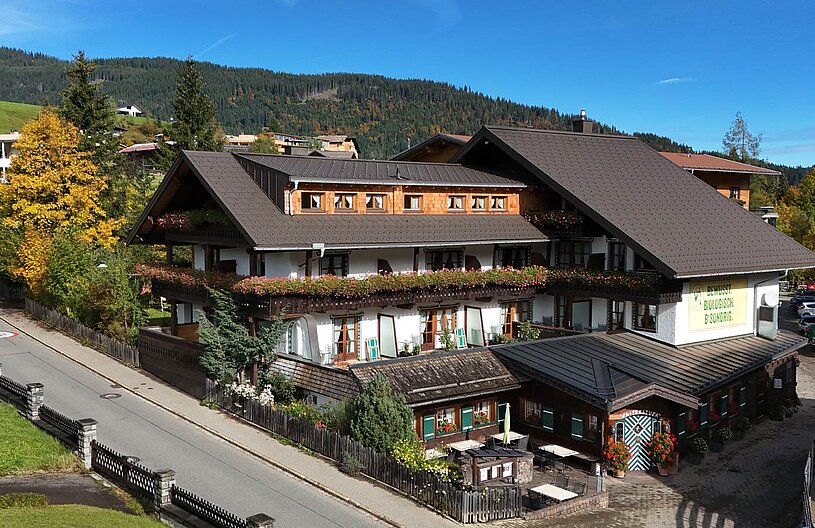 Ein malerisches Alpenhaus mit holzverkleideten Fenstern und bunten Blumenkästen. Umgeben von Bäumen und grünem Gelände, liegt es an einer ruhigen Straße. Im Vordergrund sind gedeckte Tische auf einer Terrasse zu sehen. Die Umgebung spiegelt eine idyllische Berglandschaft wider.