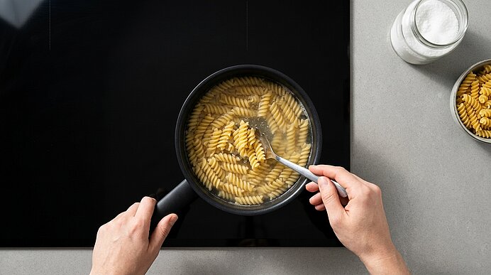 Zwei Hände rühren kochende Fusilli in einem schwarzen Stieltopf auf dem schwarzen Kochfeld um.
