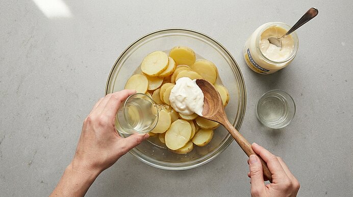 Zwei Hände bereiten Kartoffelsalat: in einer Glasschüssel liegen gekochte Kartoffelscheiben, Mayonnaise wird mit einem Holzlöffel hinzugegeben, eine Hand gießt klare Flüssigkeit aus einem kleinen Glas.