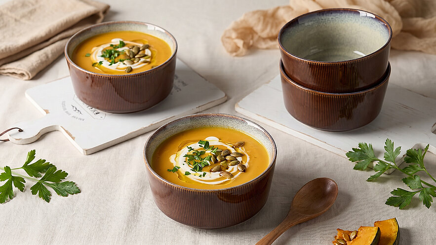 Zwei Schalen mit Kürbissuppe, garniert mit Kürbiskernen und frischen Kräutern, stehen auf einem hellen Tisch. Im Hintergrund stapeln sich weitere Schalen. Einige frische Kräuter und ein Holzlöffel liegen daneben.
