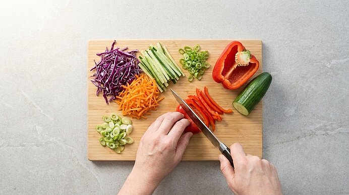 Hände schneiden rote Paprika auf einem Holzbrett; darauf liegen geschnittene Gemüsestreifen und Julienne aus Rotkohl, Gurke, Karotte und Frühlingszwiebeln.