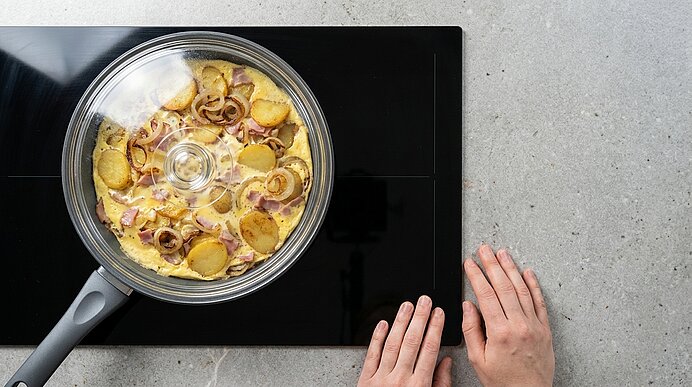 Pfanne mit Glasdeckel auf Cerankochfeld, darin goldbraune Kartoffelscheiben und Zwiebelringe; zwei Hände liegen neben dem Herd.