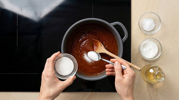 Zwei Hände geben eine weiße, granulierte Zutat aus einer kleinen Schale in einen Topf mit brauner Sauce; ein Holzlöffel liegt im Topf, daneben mehrere kleine Schälchen mit Zutaten und eine Flasche Öl.