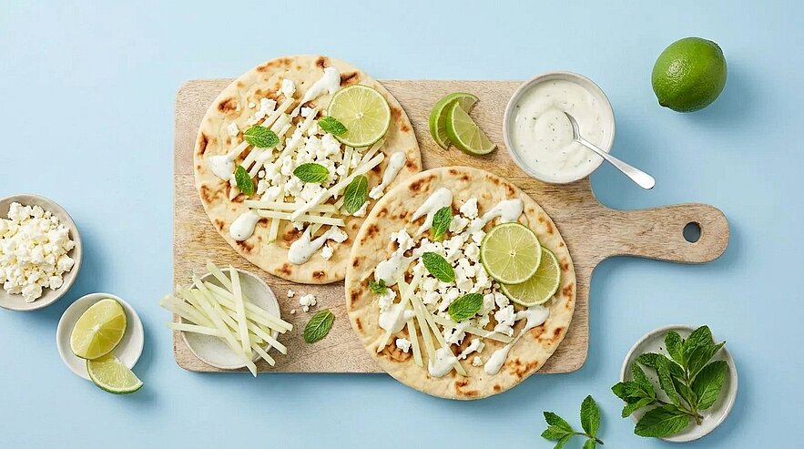 Zwei belegte Fladenbrote/Tacos auf einem Holzbrett, mit weißer Sauce, feinem Weißkraut oder Käse, Limettenscheiben und Minzblättern garniert; daneben Schälchen mit Dressing, Limettenstücken, zerbröseltem Käse und frischen Kräutern.