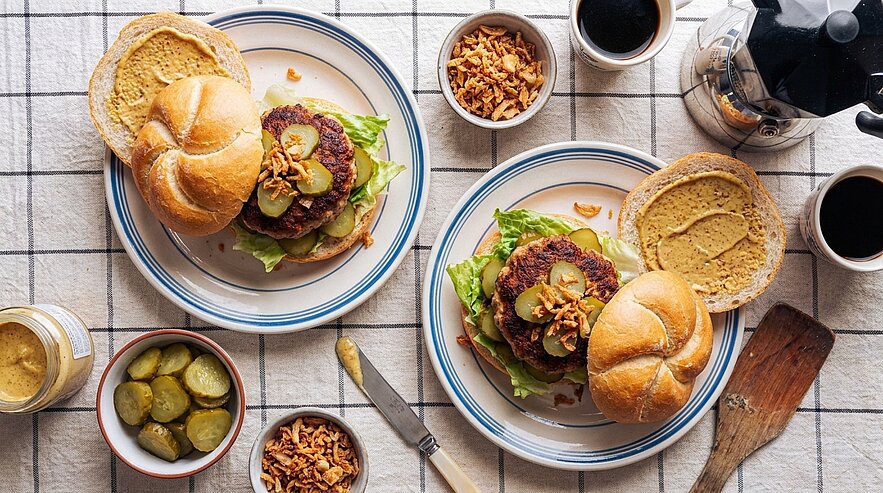 Zwei Teller mit offenen Brötchen: pflanzenbasierte Burger mit Salat und knusprigen Röstzwiebeln, daneben Schälchen mit Gewürzgurken, Senf und Kaffee.