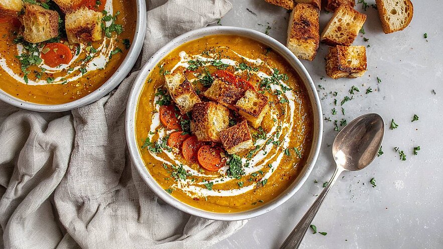 Eine cremige Karottensuppe in einer Schüssel, garniert mit geröstetem Brot, frischem Koriander und einem Schuss Sahne. Daneben liegt ein Löffel und einige unansehnliche Brotwürfel. Die Suppe hat eine leuchtend orange Farbe.