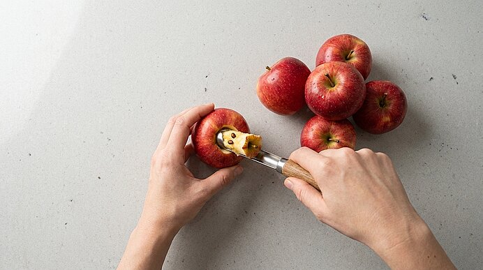 Zwei Hände entkernen mit einem Obstausstecher einen roten Apfel; mehrere rote Äpfel liegen daneben auf einer hellen Küchenarbeitsfläche.