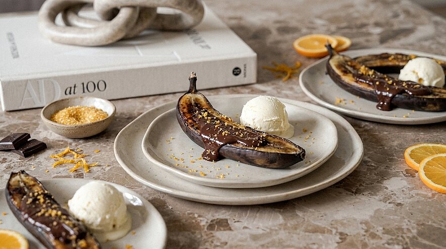 Auf Tellern gebackene, aufgeschnittene Bananen mit geschmolzener Schokolade und Streuseln, jeweils serviert mit einer Kugel Vanilleeis.