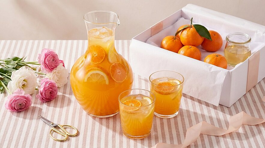 Ein Krug mit orangem Getränk, gefüllt mit Orangenscheiben, steht auf einem gestreiften Tisch. Daneben stehen zwei Gläser mit dem gleichen Getränk. Im Hintergrund ist eine Kiste mit Mandarinen und einige Blumen zu sehen.