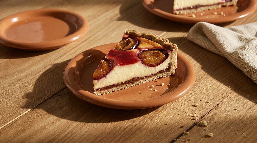 Ein Stück Käsekuchen mit Pflaumenbelag liegt auf einem rustikalen Teller. Der Kuchen ist in einer knusprigen Kruste gefasst und glänzt durch den Fruchtaufstrich. Im Hintergrund sind zwei weitere Teller und ein Geschirrtuch sichtbar.