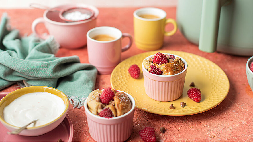 Auf dem Bild sind zwei kleine Auflaufformen mit Gebäck und frischen Himbeeren zu sehen. Daneben steht eine große Gelbe Untertasse, auf der sich eine der Formen befindet. Im Hintergrund sind Tassen und ein Tuch sichtbar. Das Gesamtbild vermittelt eine einladende Frühstücksszene.