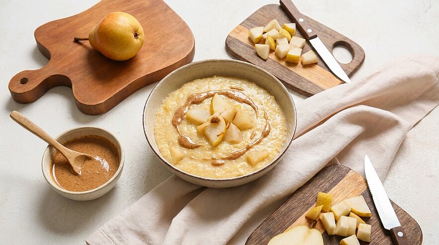 Schüssel cremiger Haferbrei, garniert mit Karamellsauce und Birnenscheiben, umgeben von geschnittenen Birnen, Schneidebrettern, Messern und einer kleinen Schale mit Zimt.