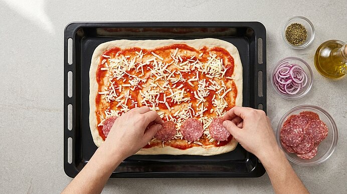 Zwei Hände legen Salamischeiben auf den mit Tomatensoße und Käse bestreuten Pizzateig auf einem Backblech.