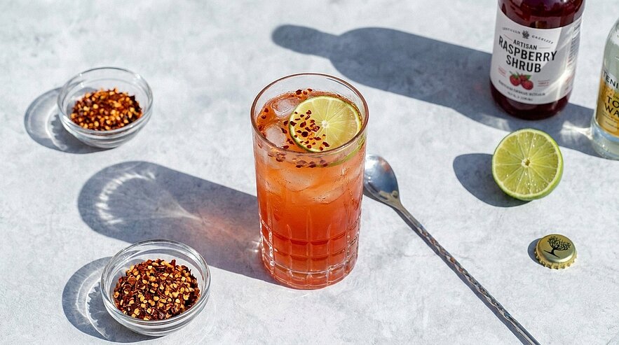 Glas mit eisgekühltem, rötlichem Cocktail, garniert mit Limettenscheibe und Chiliflocken; daneben Schälchen mit Chiliflocken, halbe Limette und Flasche Sirup.