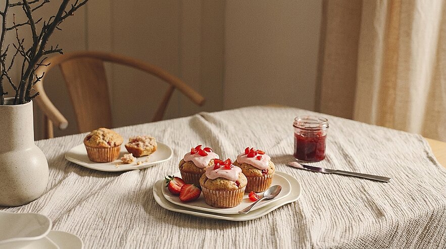 Auf dem Tisch liegen drei dekorierte Muffins mit rosa Zuckerguss und Erdbeeren. Neben ihnen steht ein weiterer Teller mit einem Muffin und ein Glas Marmelade. Im Hintergrund ist eine Vase mit einem dünnen Ast zu sehen, und die Tischdecke hat eine strukturierte Oberfläche.