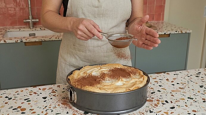 Hände einer Person in Schürze sieben Kakaopulver über eine in einer Springform stehende Torte mit Baiserhaube auf der Arbeitsplatte.
