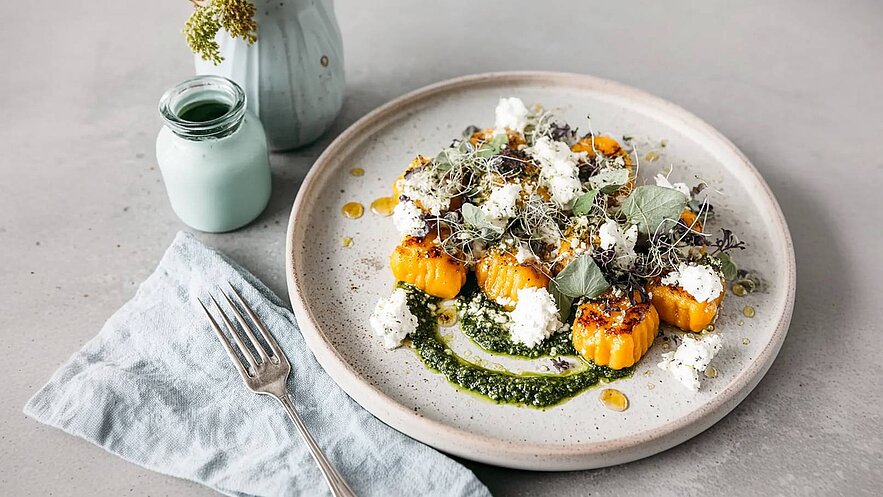 Ein Teller mit goldbraunem Gnocchi, garniert mit frischen Kräutern und kleinem Käse. Darunter befindet sich eine grüne Sauce. Neben dem Teller liegt ein blauer Servietten und eine Gabel. Im Hintergrund steht eine kleine Flasche.
