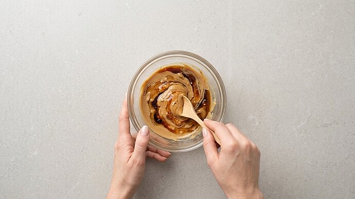 Zwei Hände verrühren Erdnussbutter und Sojasauce mit einem Holzlöffel in einer Glasschale auf hellgrauer Arbeitsfläche.