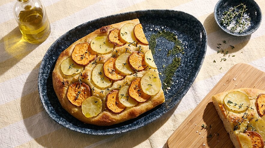 Flaches, goldbraun gebackenes Fladenbrot belegt mit dünnen Kartoffelscheiben und Kräutern, serviert auf dunklem ovalem Teller; ein Stück auf Holzbrett.