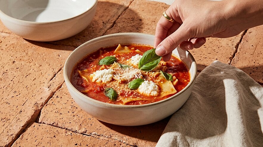 Hand legt Basilikumblätter auf eine Schüssel mit Tomaten-Lasagne-Suppe: breite Lasagneplatten in Tomatensauce, Ricotta‑Nocken und geriebener Käse.