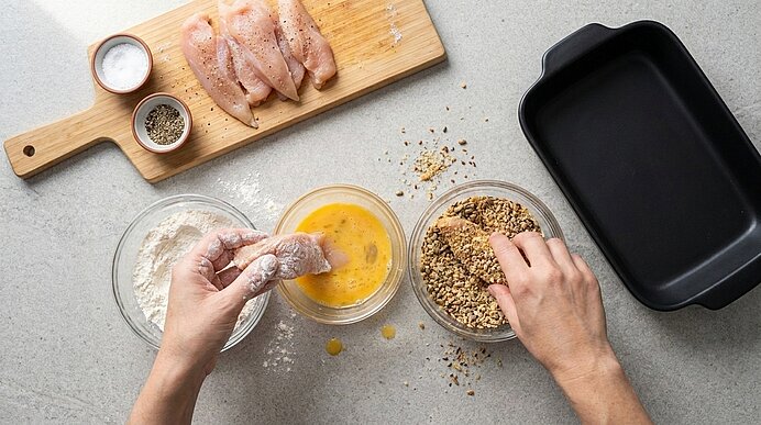Zwei Hände panieren Hähnchenbrust: rohes Fleisch auf einem Schneidebrett, Schalen mit Mehl, verquirltem Ei und grober Panade, daneben eine leere Auflaufform.