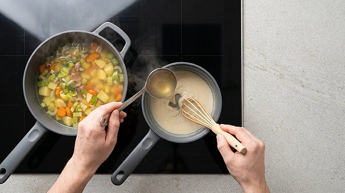 Hände über zwei Töpfen auf dem Herd: mit einem Schöpflöffel wird Brühe aus einem Gemüsetopf in einen Topf mit weißer Sauce gegossen, die rechte Hand hält einen Schneebesen.