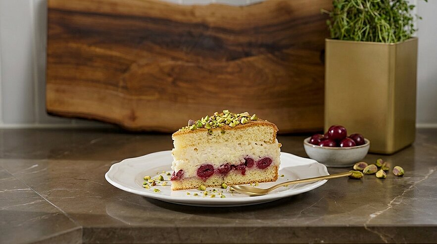 Ein Stück Kuchen steht auf einem weißen Teller. Der Kuchen hat eine cremige Schicht mit Kirschen und ist mit gehackten Pistazien dekoriert. Im Hintergrund sind Holz und ein Topf mit Kräutern sichtbar. Eine Gabel liegt neben dem Teller.