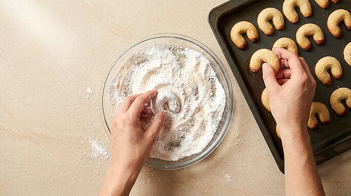 Zwei Hände tauchen ein hörnchenförmiges Vanillekipferl in eine Glasschüssel mit Puderzucker und legen bereits gezuckerte Kipferl auf ein Backblech.