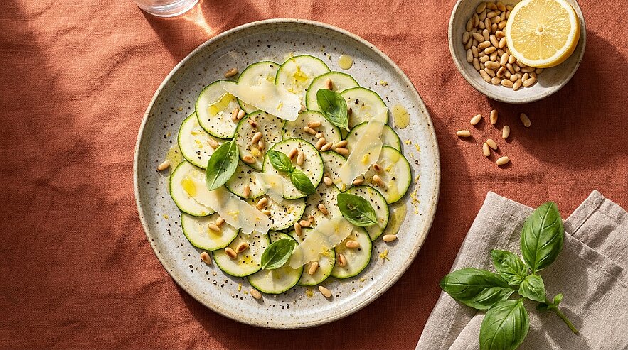 Teller mit Zucchinicarpaccio: dünne Zucchinischeiben, Basilikumblätter, gehobelter Parmesan, Pinienkerne und Olivenöl, daneben Schale mit Zitrone.