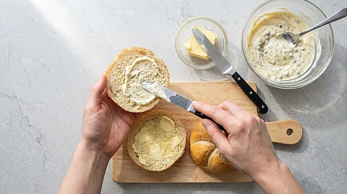 Zwei Hände bestreichen die obere Hälfte eines Brötchens mit Remoulade. Die untere Hälfte ist bereits gebuttert.