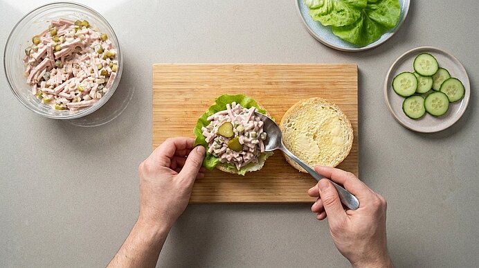 Zwei Hände häufen mit einem Löffel Fleischsalat auf eine Brötchenhälfte mit Salatblatt auf einem Holzbrett.