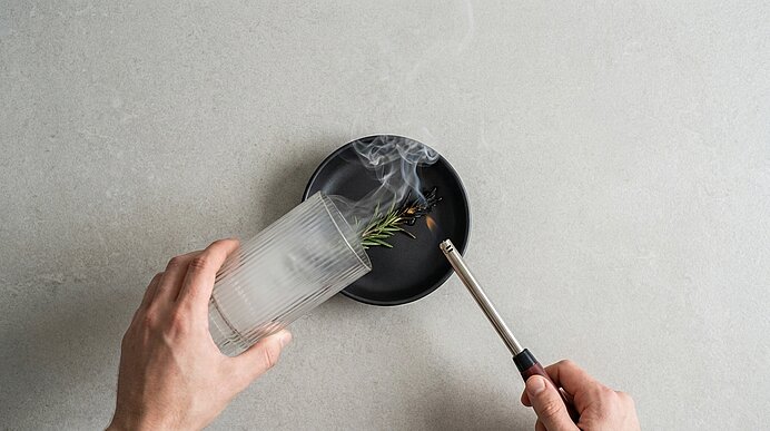 Hände setzen ein umgedrehtes Glas über einen kleinen schwarzen Teller mit Rosmarinzweig, während Rauch unter dem Glas eingeschlossen wird; die rechte Hand hält einen kleinen Brenner.