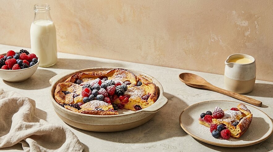 Gebratener Dutch‑Baby‑Pfannkuchen in einer flachen Keramikform, belegt mit frischen Himbeeren und Blaubeeren und bestäubt mit Puderzucker; ein Stück auf einem Teller daneben, Schale mit Beeren und Milchkanne im Hintergrund.