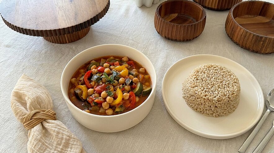 Schale mit buntem Gemüse-Kichererbsen-Eintopf (Tomaten, Paprika, Zucchini, Kräuter) neben einem Teller mit geformter Portion Vollkornreis.