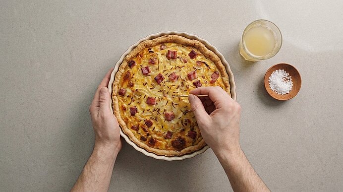 Zwei Hände halten eine herzhafte Quiche in einer Tarteform; eine Hand setzt einen Kräuterzweig als Garnitur auf die mit Speckwürfeln und Käse belegte Oberfläche.