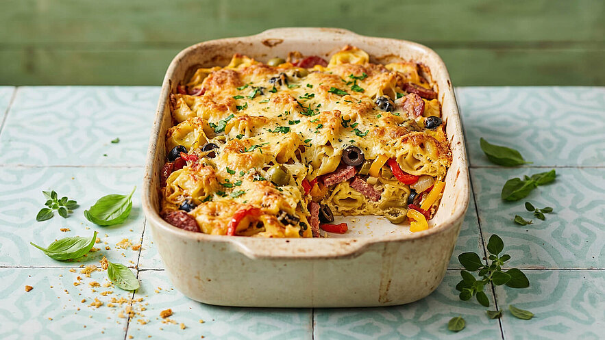 Ein Auflauf mit Nudeln, belegt mit geschmolzenem Käse, Paprika, Oliven und Schinken, serviert in einer rechteckigen Auflaufform. Frische Kräuter wie Basilikum und Oregano sind daran dekoriert. Der Hintergrund zeigt eine hellgrüne Fläche.