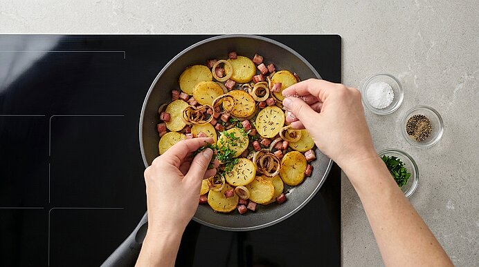 Hände bestreuen in einer Pfanne gebratene Kartoffelscheiben mit gehackter Petersilie; in der Pfanne Zwiebelringe und Wurstwürfel.