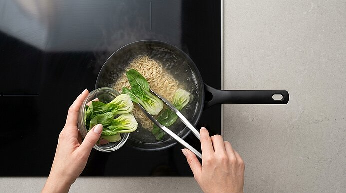 Zwei Hände geben halbierten Pak Choi mit einer Zange aus einer Schale zu den kochenden Ramen-Nudeln im Topf.