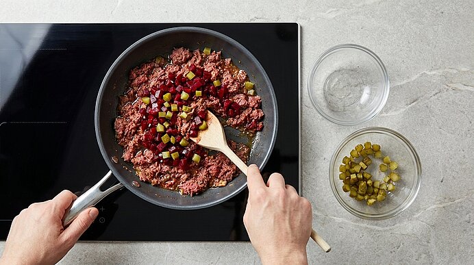 Draufsicht auf Hände, die Hackfleisch in einer Pfanne mit einem Holzlöffel rühren; gewürfelte Gewürzgurken werden hinzugegeben, zwei Glasschalen (eine mit Gurkenstücken, eine leer) stehen daneben.