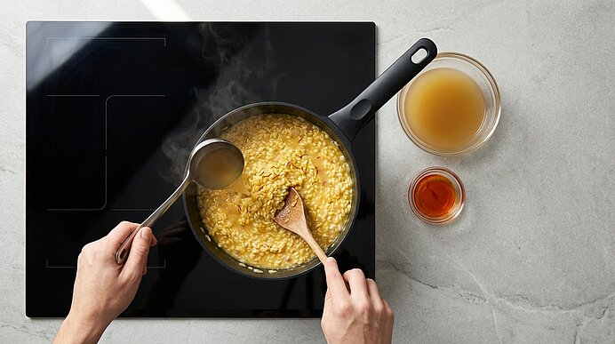 Draufsicht: Eine Person rührt cremigen Risotto in einem Topf auf dem Kochfeld mit einem Holzlöffel, während sie mit einer Kelle Brühe hinzufügt; zwei Schälchen daneben.