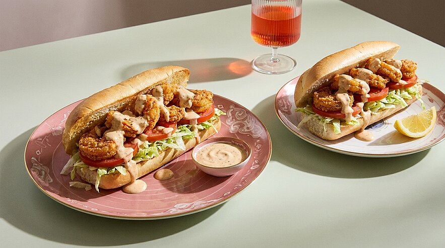 Zwei Baguette-Sandwiches mit knusprigen Garnelen, Tomaten, gehobeltem Salat und cremiger Sauce auf Tellern; dazu ein Zitronenschnitz und ein Glas mit rosafarbenem Getränk.