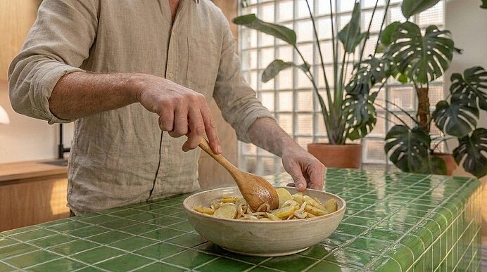 Eine Person rührt mit einem Holzlöffel in einer großen Schüssel mit geschnittenen Zutaten auf einer grün gekachelten Küchenarbeitsfläche.