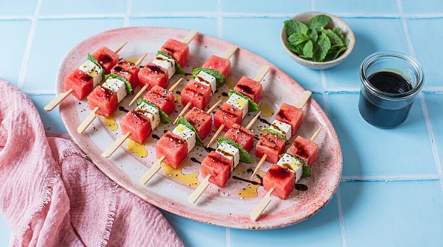 Ovaler Teller mit mehreren Spießen aus Wassermelonen- und Feta‑Würfeln, garniert mit Minzblättern und Olivenöl, serviert als erfrischende Vorspeise.